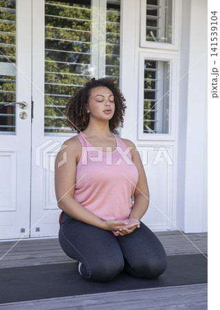 African American woman wearing pink top, leggings kneeling on yoga mat on wooden deck meditating African American woman wearing pink top, leggings kneeling on yoga mat on wooden deck meditating 141539104