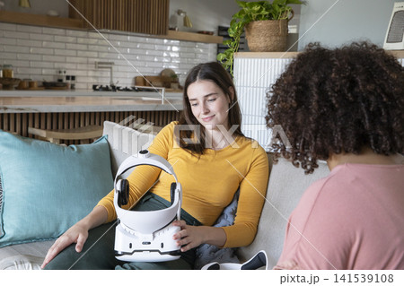 Diverse female friends testing helmet device while sitting on sofa with cushions near open kitchen Diverse female friends testing helmet device while sitting on sofa with cushions near open kitchen 141539108