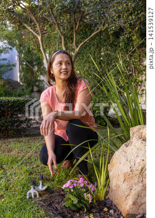 Asian woman crouching on lawn in backyard garden planting blooms using trowel and cultivator 141539127