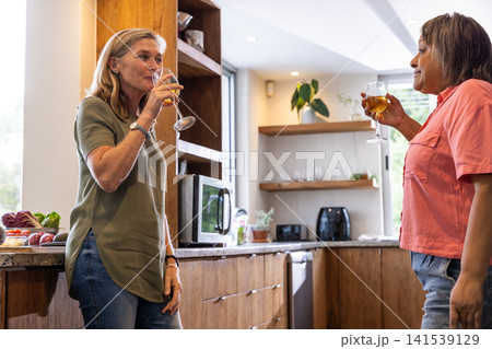 Diverse female friends clinking stemmed glasses and sipping wine on countertop in kitchen 141539129