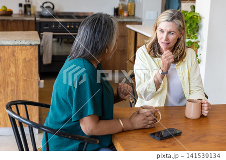 Senior female friends chatting while holding mugs at wooden kitchen table with smartphone 141539134