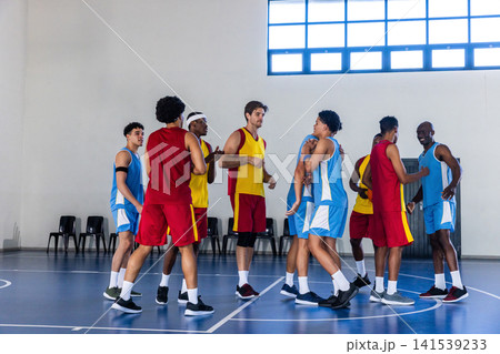 Diverse male basketball players in jerseys exchanging handshakes, hugs at gym court with chairs Diverse male basketball players in jerseys exchanging handshakes, hugs at gym court with chairs 141539233