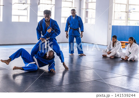 Diverse martial arts class grappling, throwing in blue gis at dojo on mats, wooden boards Diverse martial arts class grappling, throwing in blue gis at dojo on mats, wooden boards 141539244