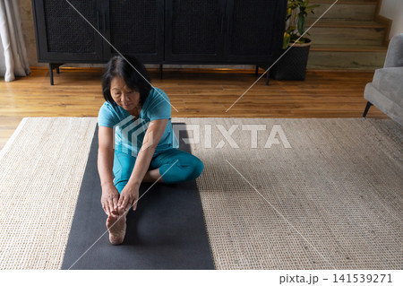 Senior Asian woman stretching in turquoise activewear on black yoga mat in living room, copy space 141539271