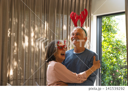 Senior couple laughing and hugging in living room wearing red antler headbands and reindeer glasses 141539292