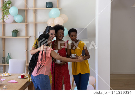 Diverse female friends hugging, posing for selfie with phone by dining table, cupcakes, coffee mugs 141539354