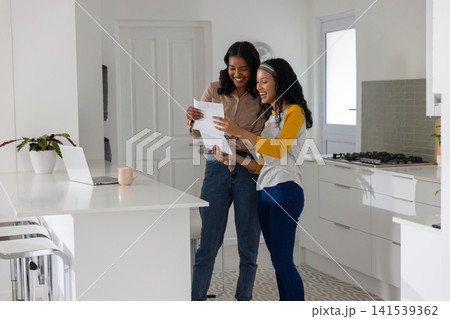 Diverse mom and teenage daughter reviewing papers at white island with laptop and mug Diverse mom and teenage daughter reviewing papers at white island with laptop and mug 141539362