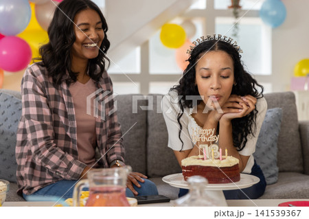 Diverse mom and teenage daughter blowing out candles on 18th birthday cake at home, copy space 141539367