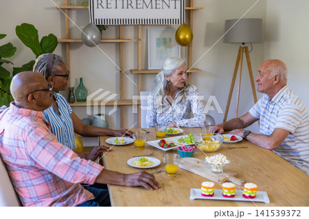 Diverse senior friends chatting at home dining table with fruit, juice under retirement banner Diverse senior friends chatting at home dining table with fruit, juice under retirement banner 141539372