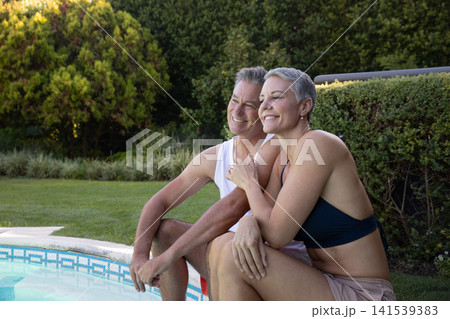 Senior couple in swimwear sitting on pool edge in backyard by water, mosaic tile border Senior couple in swimwear sitting on pool edge in backyard by water, mosaic tile border 141539383