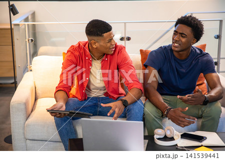 Diverse male friends sitting on beige sofa in modern living room, using tablet and laptop 141539441