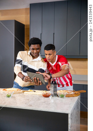 Diverse male friends preparing pizzas at marble kitchen island using tablet and wooden pizza boards 141539443