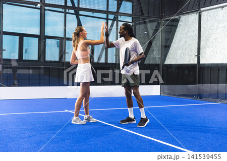 Diverse teammates standing facing each other near net on blue padel court indoors holding rackets 141539455
