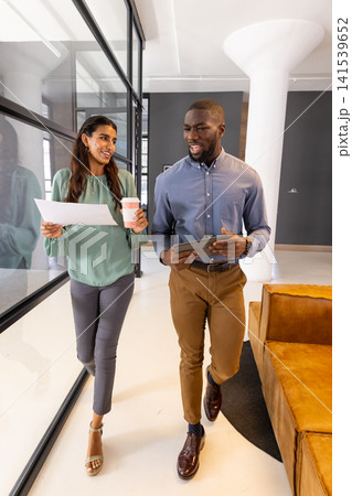 Diverse coworkers walking office corridor carrying document, coffee cup, tablet by column Diverse coworkers walking office corridor carrying document, coffee cup, tablet by column 141539652