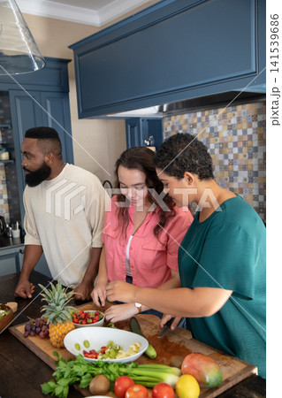 Diverse friends preparing fruit salad at kitchen with large cutting board and mixing bowl Diverse friends preparing fruit salad at kitchen with large cutting board and mixing bowl 141539686