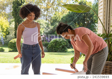 Diverse mother and daughter painting wooden planks on porch with pink paint and brushes Diverse mother and daughter painting wooden planks on porch with pink paint and brushes 141539693