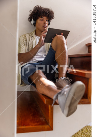 African american man sitting on staircase at home using tablet with wireless headphones African american man sitting on staircase at home using tablet with wireless headphones 141539714