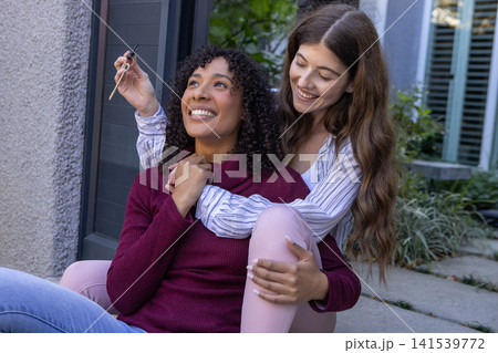Female partners sitting on porch steps by potted plants, wrapping arms, lifting dropper bottle Female partners sitting on porch steps by potted plants, wrapping arms, lifting dropper bottle 141539772