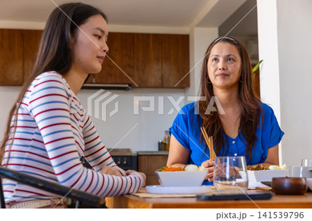 Asian mother and daughter sitting at dining table in kitchen holding chopsticks over bowl Asian mother and daughter sitting at dining table in kitchen holding chopsticks over bowl 141539796