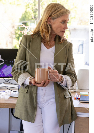 Middle-aged woman wearing blazer holding mug near desk at office displaying chart on monitor Middle-aged woman wearing blazer holding mug near desk at office displaying chart on monitor 141539809
