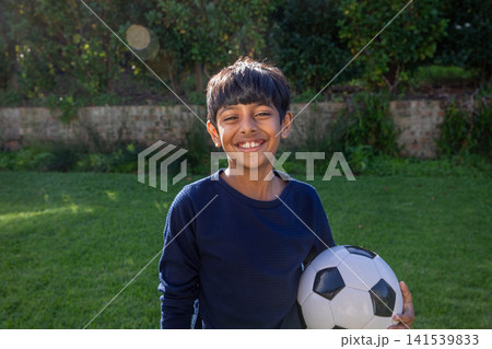 Asian boy in navy blue shirt smiling and holding soccer ball in lush green backyard Asian boy in navy blue shirt smiling and holding soccer ball in lush green backyard 141539833