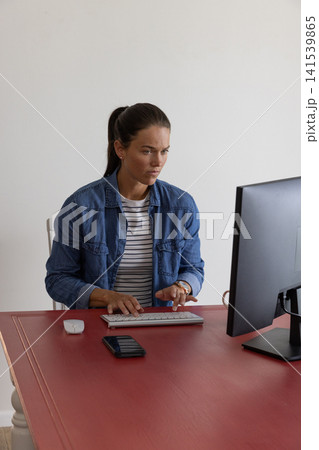 White woman sitting at desk at home typing on keyboard, checking computer monitor by smartphone White woman sitting at desk at home typing on keyboard, checking computer monitor by smartphone 141539865