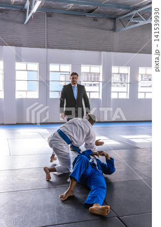Diverse male martial artists in gis grappling in dojo while referee in suit watching Diverse male martial artists in gis grappling in dojo while referee in suit watching 141539930