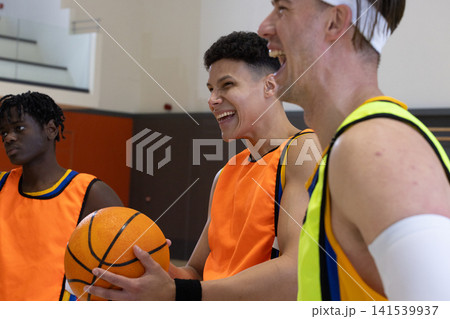 Diverse male friends smiling and holding basketball while chatting on court wearing jerseys Diverse male friends smiling and holding basketball while chatting on court wearing jerseys 141539937