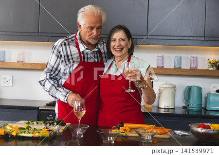 Senior couple wearing red aprons preparing vegetables at modern kitchen island with wine glasses Senior couple wearing red aprons preparing vegetables at modern kitchen island with wine glasses 141539971