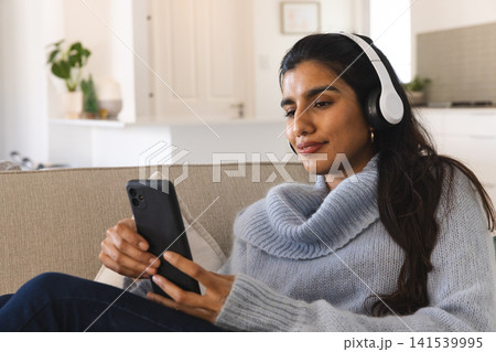 South Asian woman sitting on couch at home scrolling smartphone wearing headphones, copy space South Asian woman sitting on couch at home scrolling smartphone wearing headphones, copy space 141539995