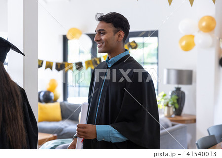 Asian friends celebrating graduation inside decorated living room with gowns balloons, diploma 141540013