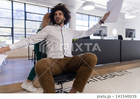 Diverse coworkers pushing rolling office chair and holding paper document in office interior Diverse coworkers pushing rolling office chair and holding paper document in office interior 141540039