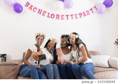 Diverse female friends taking smartphone selfie on sofa at home under bachelorette party banner Diverse female friends taking smartphone selfie on sofa at home under bachelorette party banner 141540044