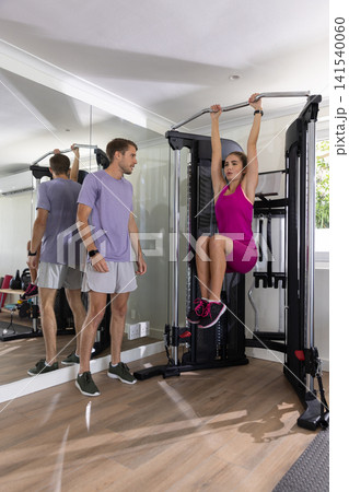 Male trainer guiding female client in pink performing knee-raises on bar in home gym Male trainer guiding female client in pink performing knee-raises on bar in home gym 141540060