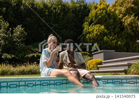 Diverse couple sitting on tiled pool edge dipping legs in water and sipping drinks in shades 141540075