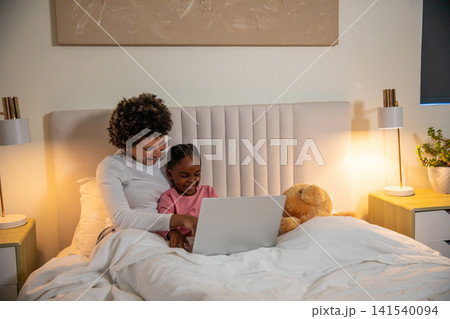 Diverse mother with daughter smiling while using laptop with teddy bear on bed with headboard Diverse mother with daughter smiling while using laptop with teddy bear on bed with headboard 141540094