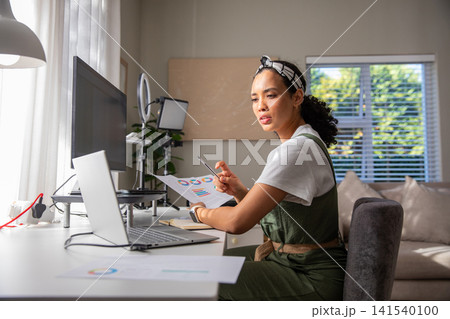 Woman holding printed chart, pen while studying data on laptop at home office corner Woman holding printed chart, pen while studying data on laptop at home office corner 141540100