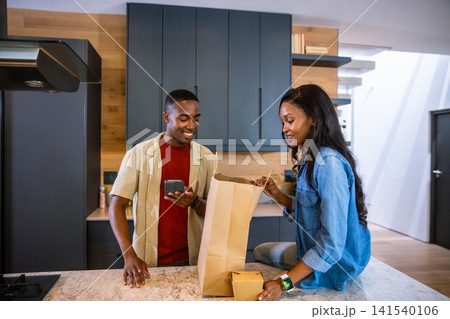 African American couple unpacking groceries from bag at kitchen island while checking smartphone 141540106