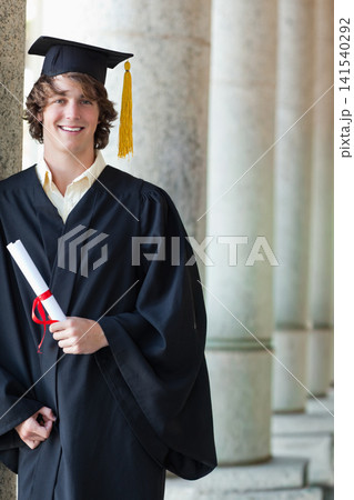 Male graduate leaning on campus column holding diploma wearing gown and cap with yellow tassel Male graduate leaning on campus column holding diploma wearing gown and cap with yellow tassel 141540292