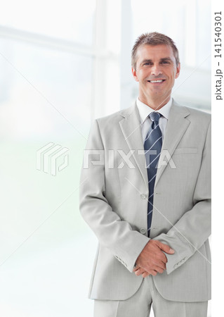 Mature man standing right in glass lobby, clasping hands, wearing gray suit, navy tie, copy space Mature man standing right in glass lobby, clasping hands, wearing gray suit, navy tie, copy space 141540301