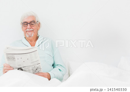 Senior man sitting in bed reading broadsheet newspaper wearing mint pajamas, glasses, copy space 141540330