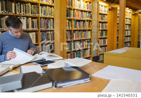 Middle-aged man sitting at wooden study table in library turning open book pages, copy space Middle-aged man sitting at wooden study table in library turning open book pages, copy space 141540331