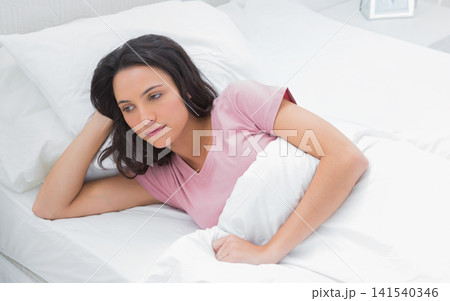 Female reclining on bed in bedroom, holding white duvet, resting on pillows, wearing pink T-shirt 141540346