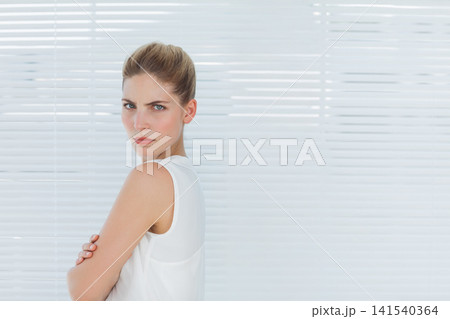 Mid 20s woman standing arms crossed, wearing white tank top before horizontal blinds, copy space 141540364