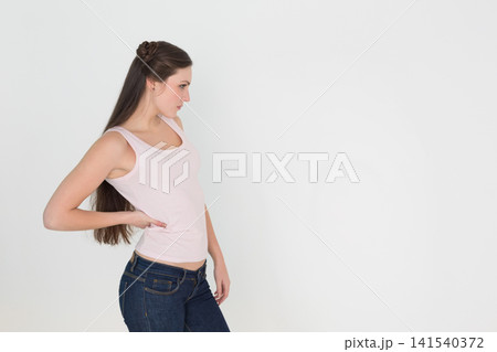 Female model posing profile left white backdrop wearing pink tank, jeans, stud earrings, copy space Female model posing profile left white backdrop wearing pink tank, jeans, stud earrings, copy space 141540372
