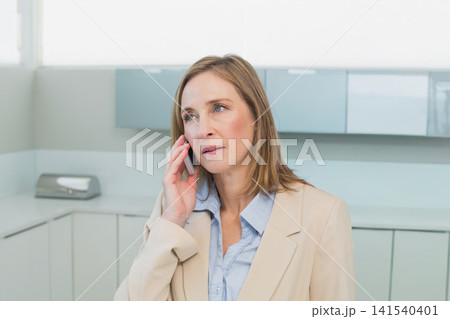 Mature woman in beige blazer holding smartphone, gazing left in small clinic with tissue dispenser Mature woman in beige blazer holding smartphone, gazing left in small clinic with tissue dispenser 141540401