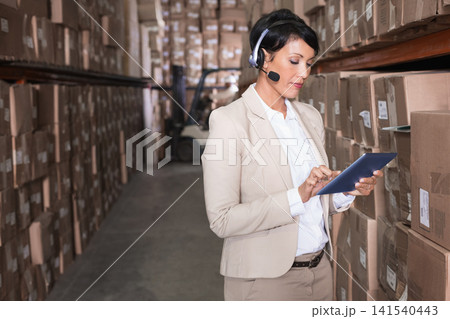 Mature adult woman wearing beige suit using tablet and headset in warehouse aisle with forklift Mature adult woman wearing beige suit using tablet and headset in warehouse aisle with forklift 141540443