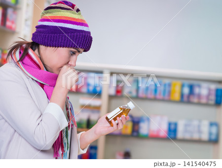 Female shopper in pharmacy examining amber bottle covering mouth wearing hat and scarf, copy space Female shopper in pharmacy examining amber bottle covering mouth wearing hat and scarf, copy space 141540475