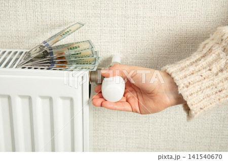 Heating season. Hand adjusts thermostat while money rests on radiator. Cost management concept Heating season. Hand adjusts thermostat while money rests on radiator. Cost management concept 141540670