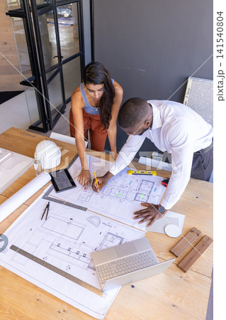 Diverse coworkers leaning over blueprints at office table adjusting models using tablet and laptop Diverse coworkers leaning over blueprints at office table adjusting models using tablet and laptop 141540804